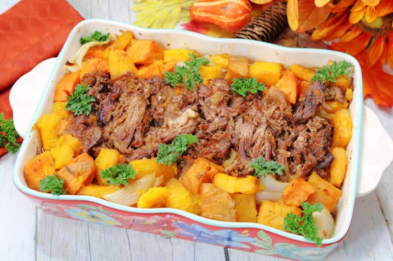 A hearty dish of tender shredded beef surrounded by roasted butternut squash and garnished with fresh parsley in a floral baking dish.